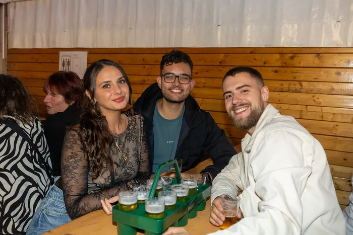 Drei lächelnde junge Menschen sitzen an einem Tisch mit Biergläsern bei der Q-Stall-Party.