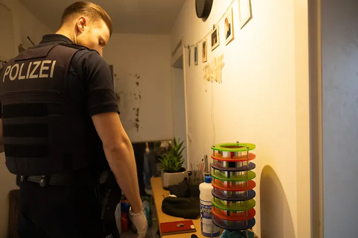 Ein Polizist mit Schutzweste steht im Hintergrund eines Raumes, in dem verschiedene Rollen bunter Klebebänder und Flüssigkeiten auf einem Tisch stehen.