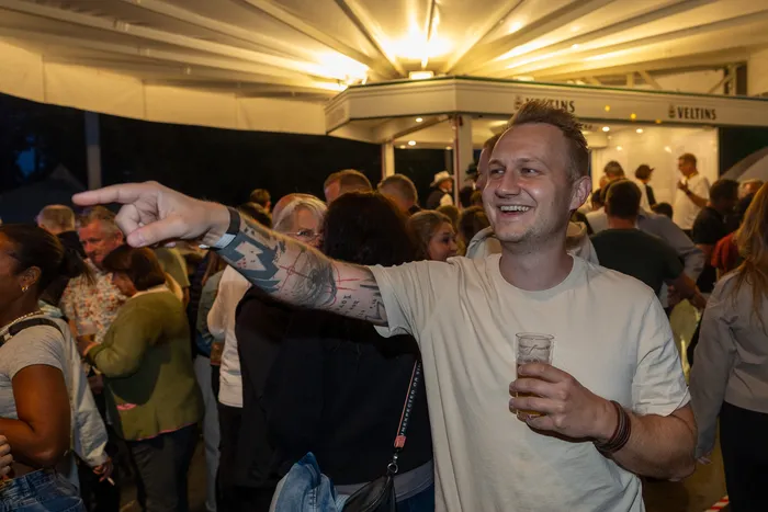 Ein lächelnder Mann mit Tätowierung am Arm zeigt bei einer Party in eine Richtung und hält ein Glas Bier.