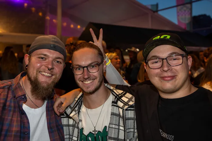 Drei lächelnde Männer, einer davon mit einem Peace-Zeichen, posieren bei der zehnten Q-Stall-Party in Schalksmühle.