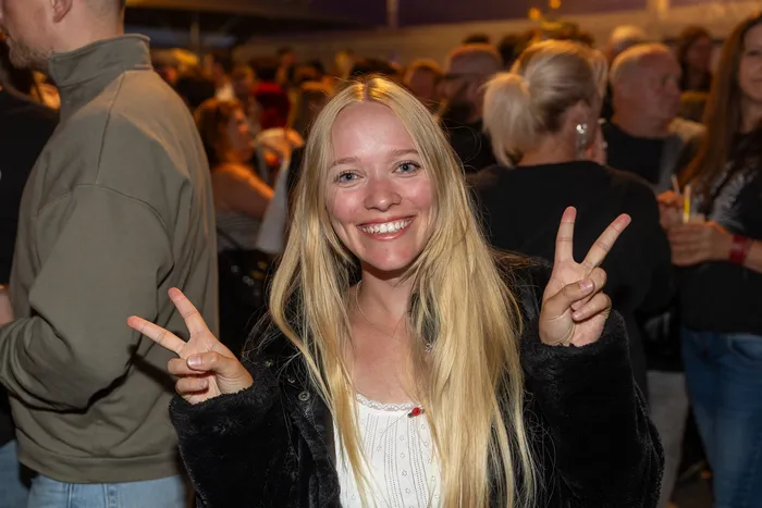Eine lächelnde junge Frau mit langen blonden Haaren zeigt mit beiden Händen das Peace-Zeichen vor einer Menschenmenge.