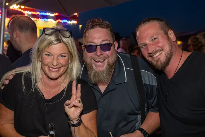 Eine blonde Frau mit Peace-Zeichen und zwei bärtige Männer lächeln bei einer Party.