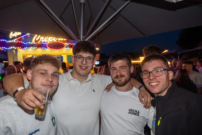 Vier junge Männer posieren lächelnd bei einer Party vor einem hell erleuchteten Crepes-Stand.