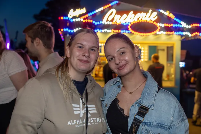 Zwei lächelnde junge Frauen stehen vor einem hell erleuchteten Creperie-Stand auf einer Party.