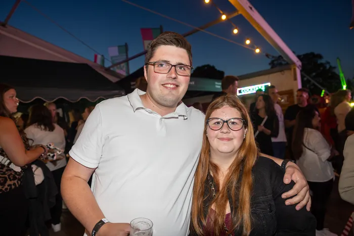 Ein junger Mann in einem weißen Poloshirt und eine junge Frau mit Brille lächeln gemeinsam bei einer Abendveranstaltung.