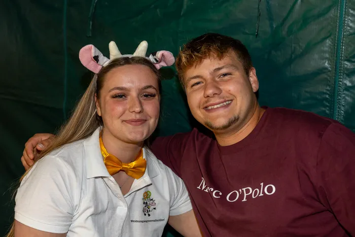 Eine lächelnde junge Frau mit Kuhohren und gelber Fliege sowie ein lächelnder junger Mann in einem weinroten T-Shirt.