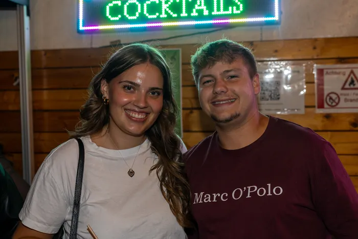 Eine junge Frau und ein junger Mann lächeln vor einer Holzwand mit einem neonfarbenen „COCKTAILS“-Schild.