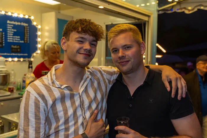 Zwei lächelnde junge Männer posieren vor einem Verkaufsstand bei der Q-Stall-Party in Schalksmühle.