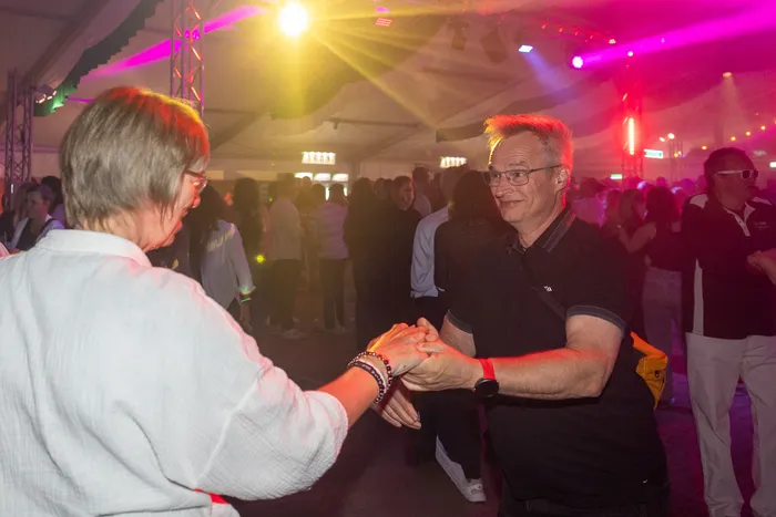 Ein Mann in einem schwarzen Poloshirt und eine Frau in Weiß tanzen Hand in Hand auf einer Party mit farbenfroher Beleuchtung.