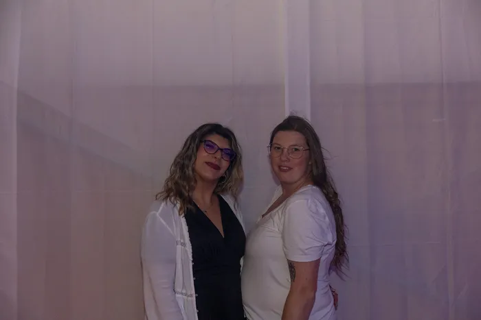Zwei Frauen mit Brillen posieren lächelnd eng beieinander vor einem weißen Vorhang.