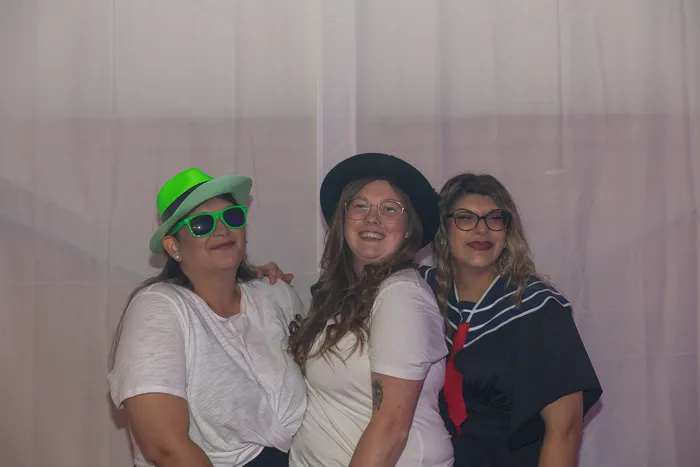 Drei lächelnde Frauen posieren vor einem weißen Hintergrund; eine trägt einen neongrünen Hut und eine grüne Sonnenbrille, eine andere einen schwarzen Hut.