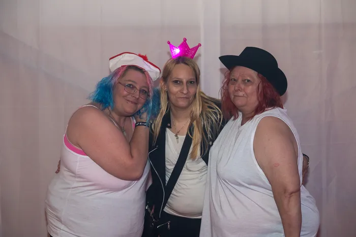 Drei Frauen mit Party-Accessoires wie einer leuchtenden rosa Krone, einem rot-weißen Hut und einem schwarzen Hut posieren gemeinsam.