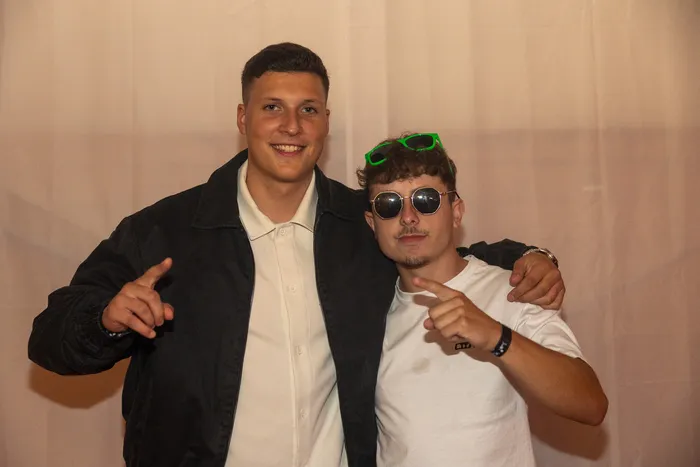 Zwei junge Männer posieren lächelnd vor einem hellen Hintergrund auf der Q-Stall-Party.