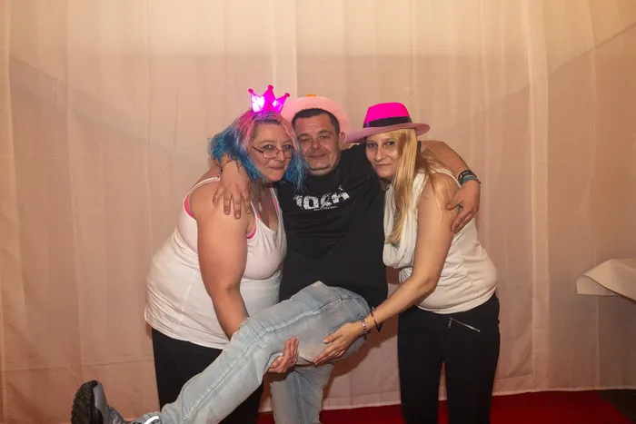 Drei Gäste mit rosa Party-Accessoires posieren gemeinsam vor einem weißen Vorhang.