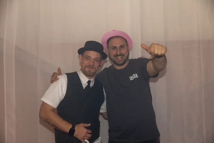 Zwei Männer posieren vor einem weißen Vorhang, einer mit schwarzem Hut und Weste, der andere mit rosa Hut und einem Daumen nach oben.
