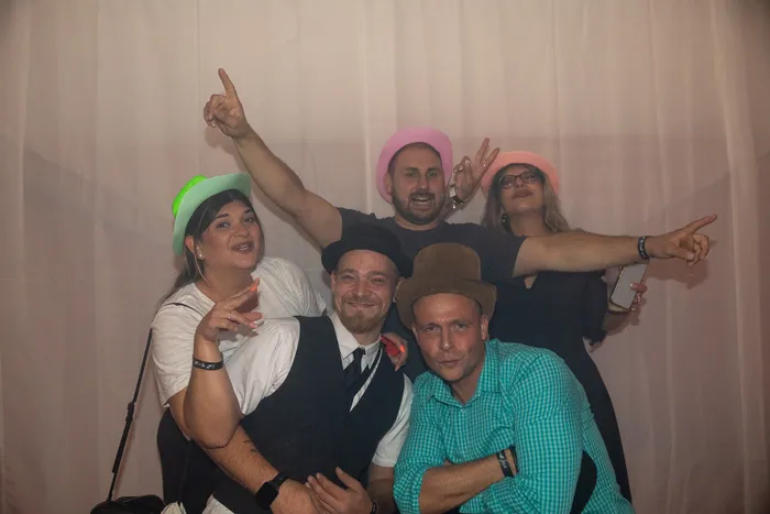 Fünf fröhliche Partygäste mit bunten Hüten posieren für ein Gruppenfoto vor einem hellen Vorhang.