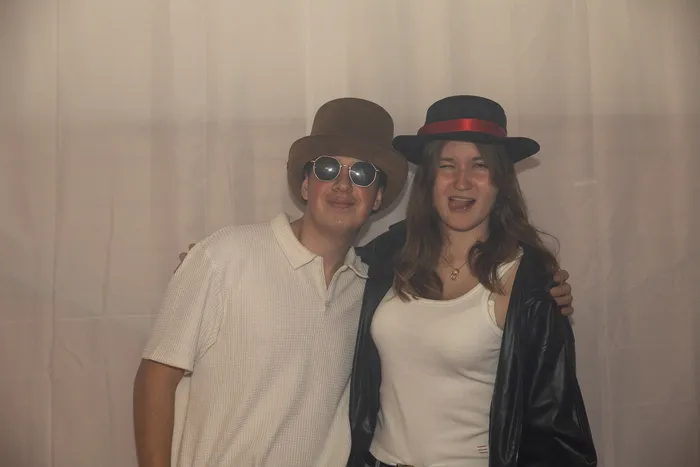 Zwei junge Menschen posieren bei einer Party; einer trägt einen braunen Hut und eine Sonnenbrille, die andere einen schwarzen Hut und eine Lederjacke.