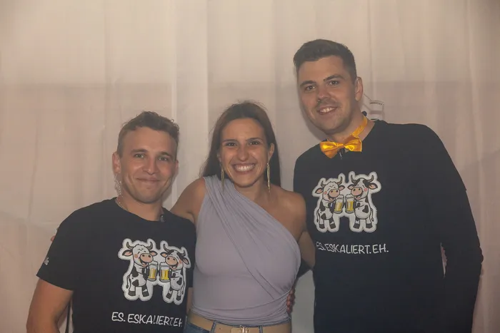 Drei lächelnde Personen posieren vor einem weißen Hintergrund, zwei Männer tragen schwarze T-Shirts mit einem Kuh-Motiv und dem Text „ES.ESKALIERTEH.“. 