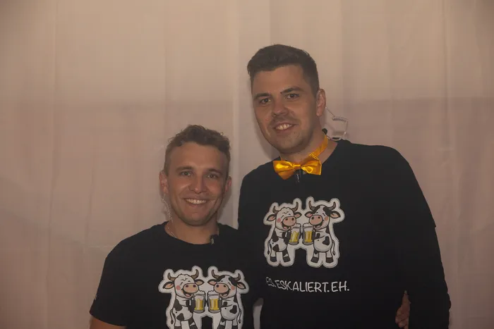 Zwei lächelnde Männer in schwarzen T-Shirts mit einem Kuh-Motiv und dem Schriftzug „ESKALIERT.EH.“, einer trägt eine gelbe Fliege.
