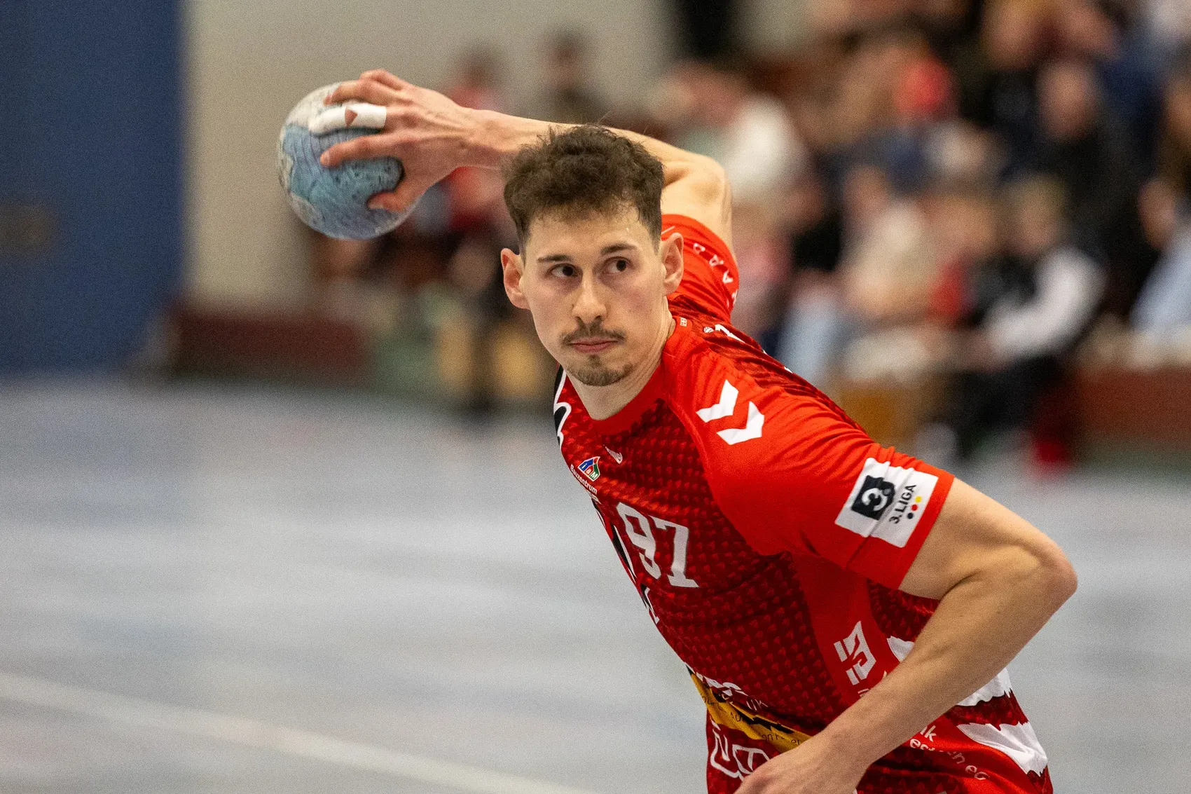 Handballspieler wirft den Ball während eines Spiels. Er trägt ein rotes Trikot mit der Nummer 87.
