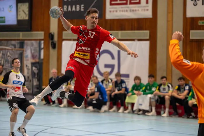 Handballspieler in rotem Trikot springt während eines Spiels in der Luft, um den Ball zu werfen. Im Hintergrund weitere Spieler und Zuschauer in einer Sporthalle.