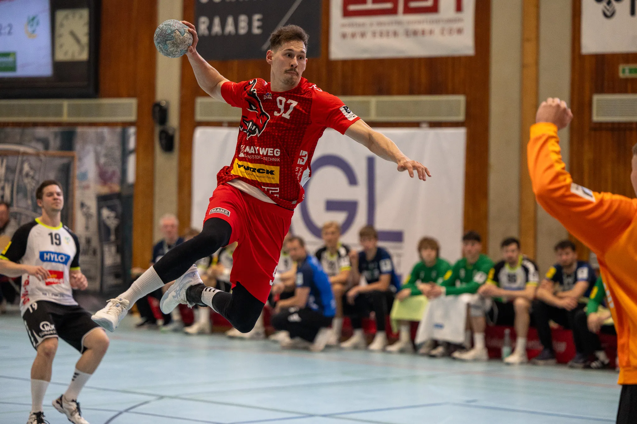 Handballspieler in rotem Trikot springt während eines Spiels in der Halle zum Wurf ab. Im Hintergrund weitere Spieler und Zuschauer.