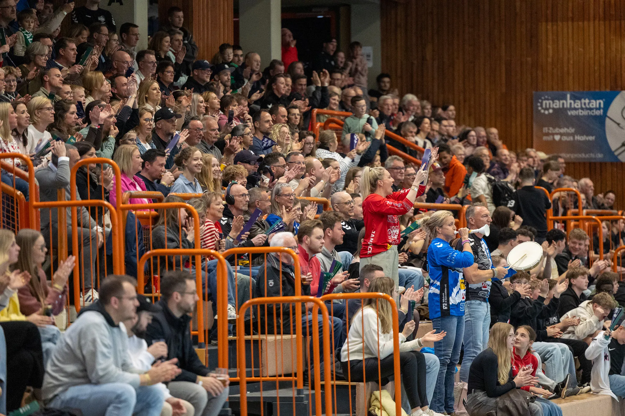 Zuschauer in einer Sporthalle klatschen und jubeln bei einem Spiel der SGSH Dragons, während im Hintergrund eine Werbetafel für Manhattan zu sehen ist.