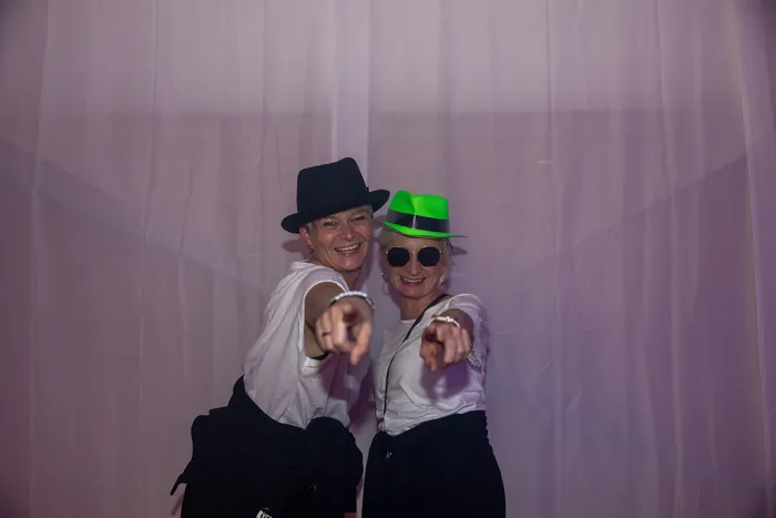 Zwei lächelnde Frauen mit einem schwarzen und einem neongrünen Hut zeigen mit den Fingern in die Kamera.