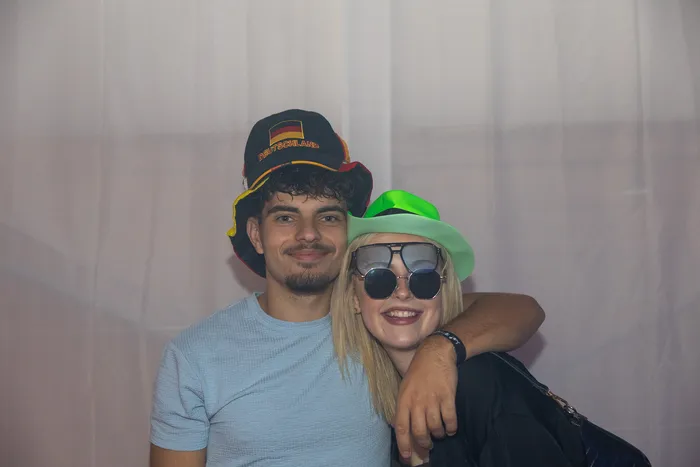 Ein lächelndes Paar posiert bei einer Party; der Mann trägt einen Hut mit der Aufschrift „Deutschland“ und die Frau einen grünen Hut sowie eine Sonnenbrille.