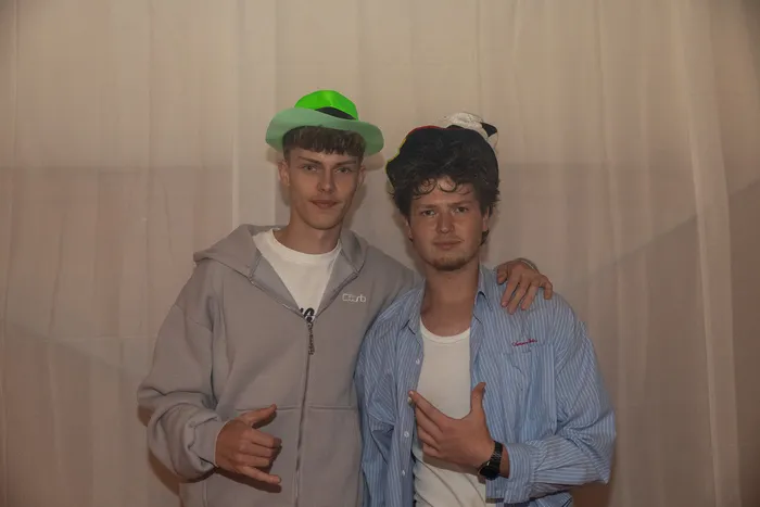 Zwei junge Männer mit Hüten posieren mit Handgesten vor einem weißen Vorhang bei der Q-Stall-Party.
