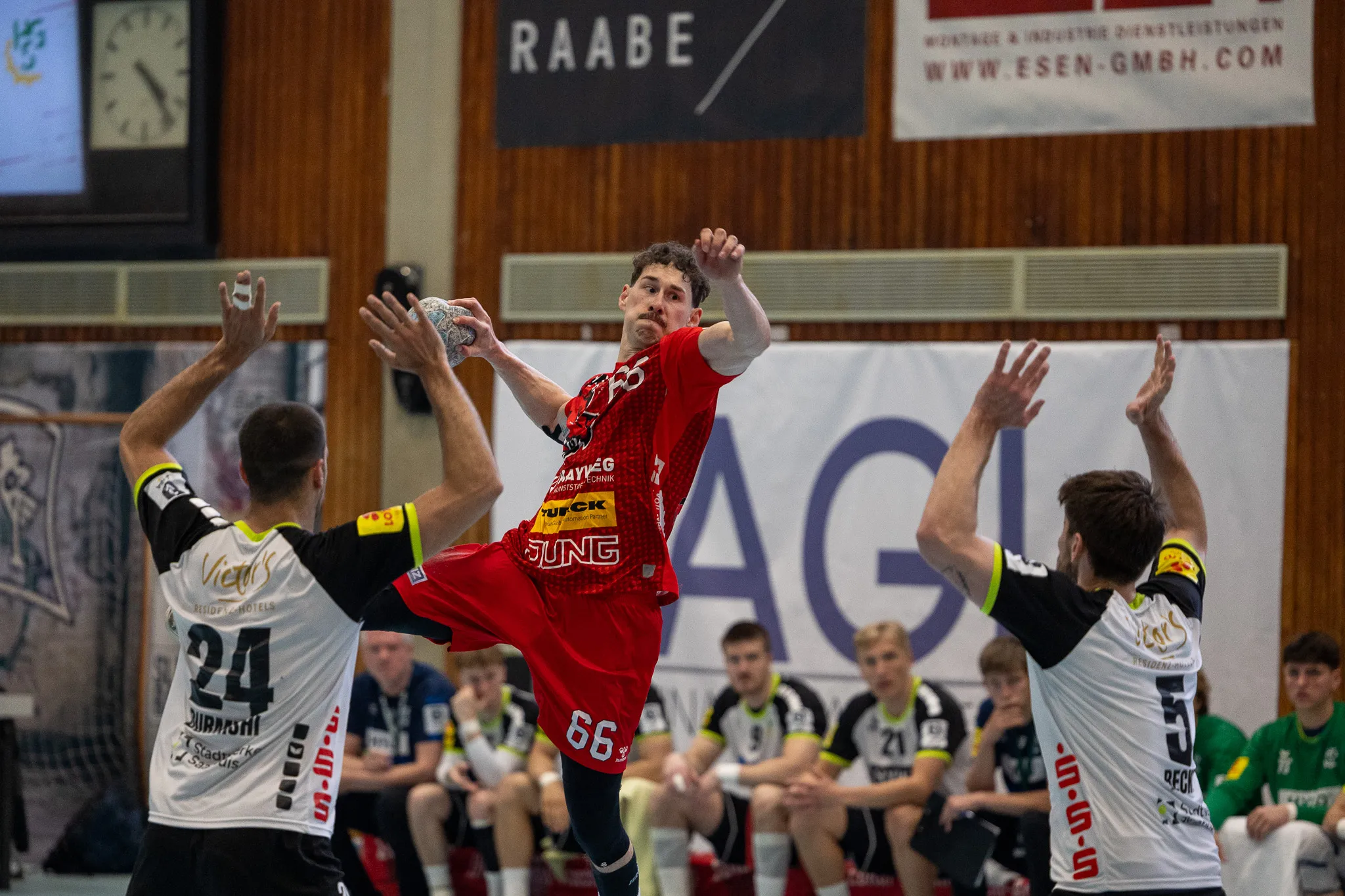Ein Handballspieler in rotem Trikot springt hoch, um den Ball zu werfen, umgeben von Gegenspielern in schwarzen und gelben Trikots während eines Spiels in einer Sporthalle.