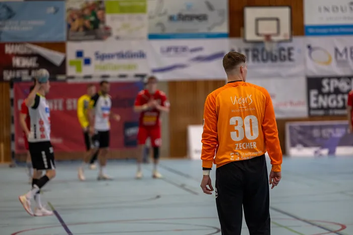 Handballspieler mit der Rückennummer 30 geht vom Betrachter weg, während im Hintergrund weitere Spieler in einer Halle spielen.