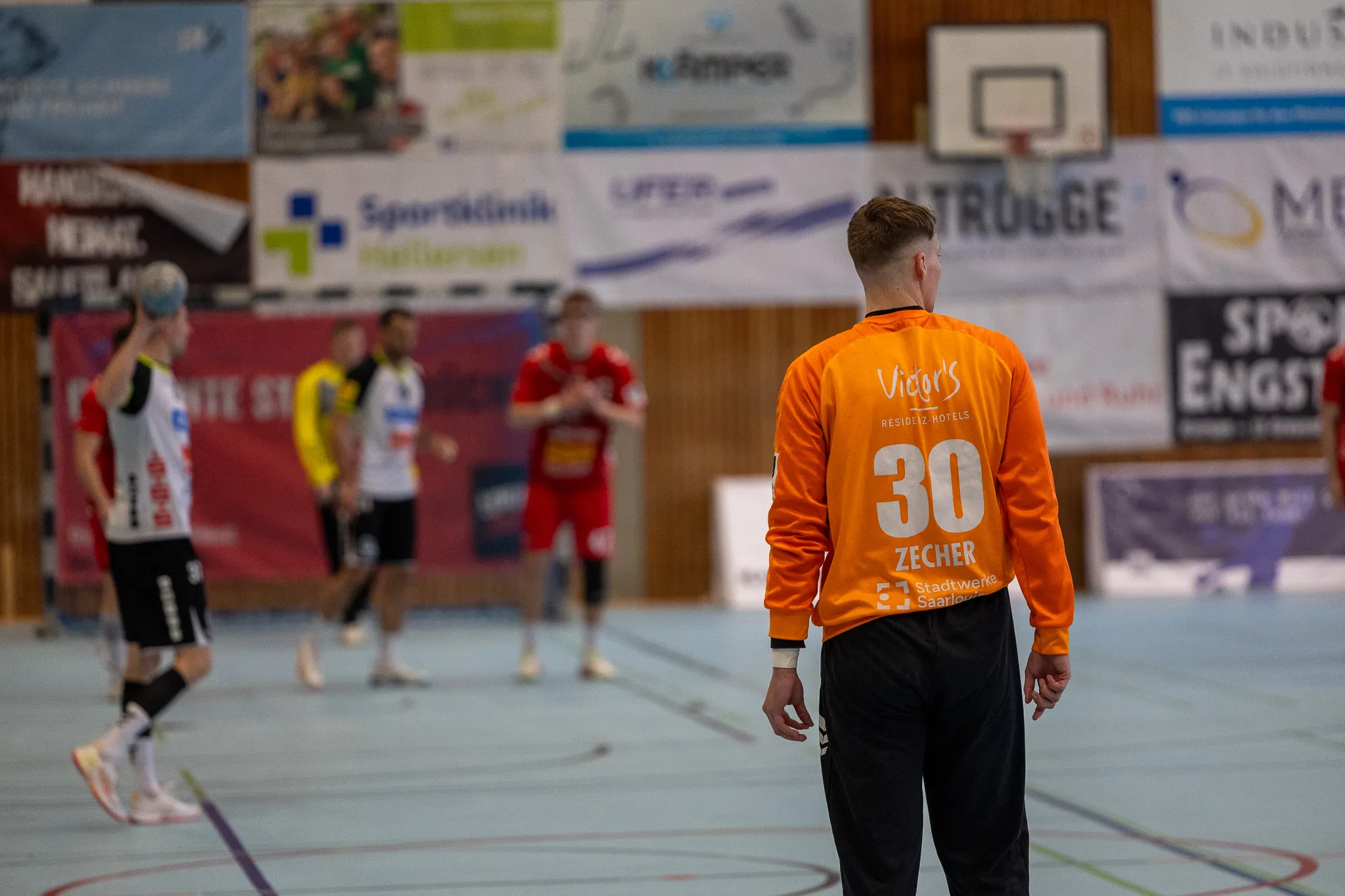 Handballspieler mit der Rückennummer 30 geht vom Betrachter weg, während im Hintergrund weitere Spieler in einer Halle spielen.