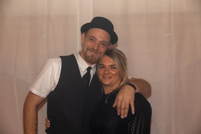 Ein Mann mit schwarzem Hut, Weste und Krawatte umarmt eine lächelnde Frau mit blonden Haaren vor einem hellen Vorhang.