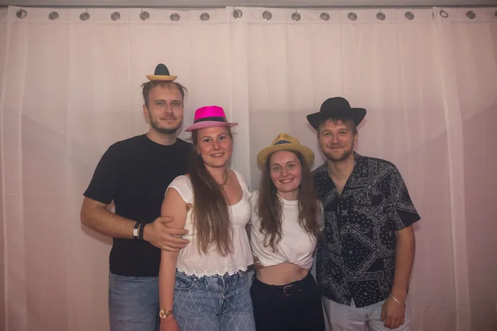 Vier junge Menschen mit bunten Partyhüten posieren lächelnd vor einem weißen Vorhang.