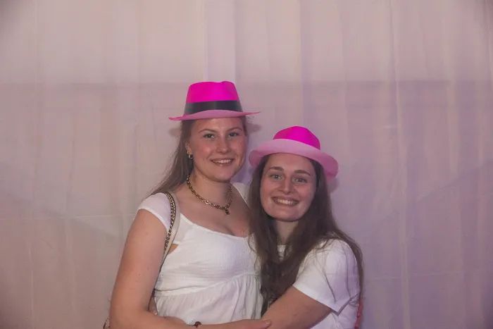 Zwei lächelnde junge Frauen mit pinkfarbenen Hüten und weißen Oberteilen posieren vor einem hellen Hintergrund.