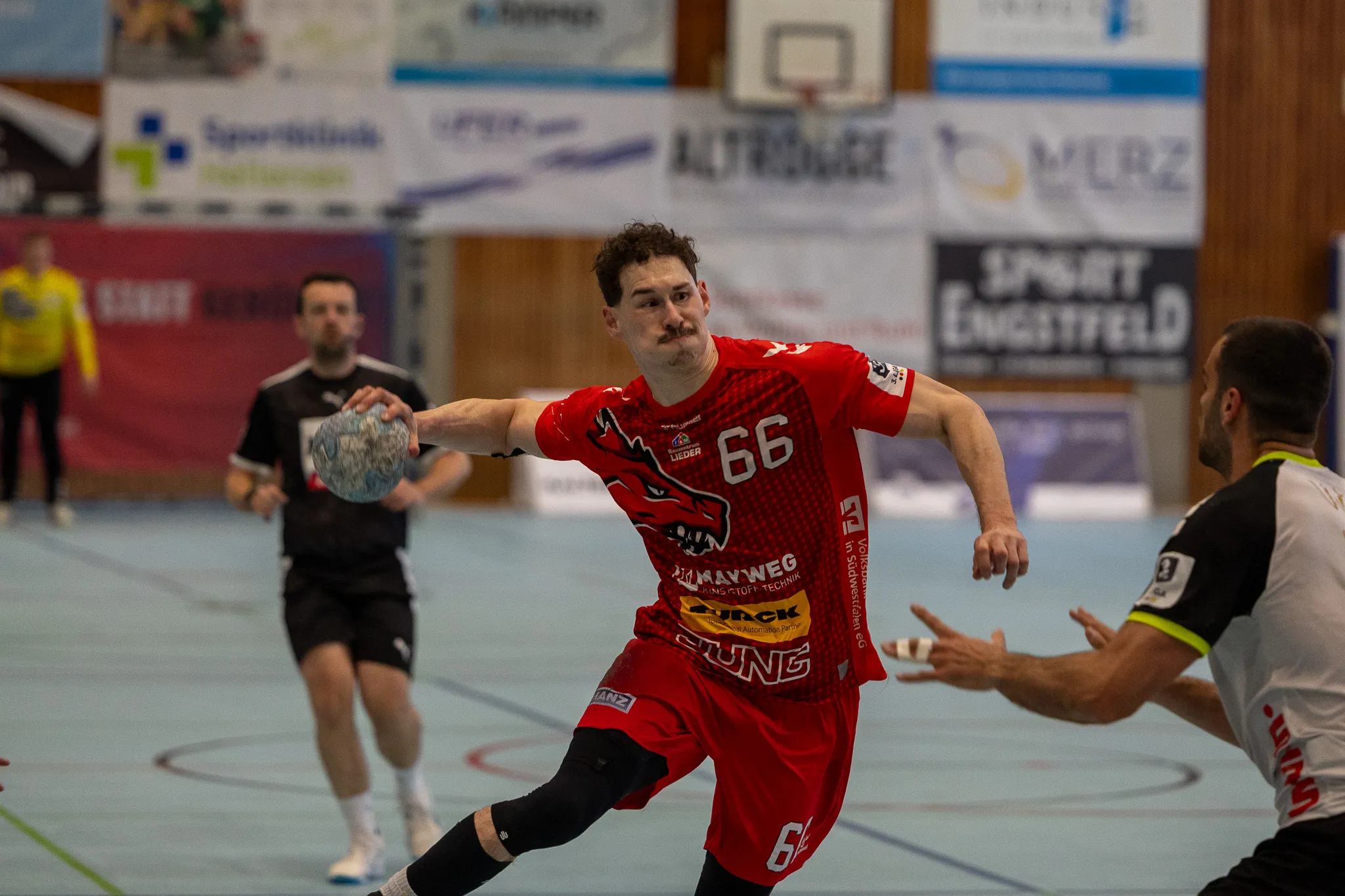 Handballspieler in rotem Trikot mit der Nummer 66 wirft den Ball während eines Spiels. Im Hintergrund sind weitere Spieler und Zuschauer zu sehen.