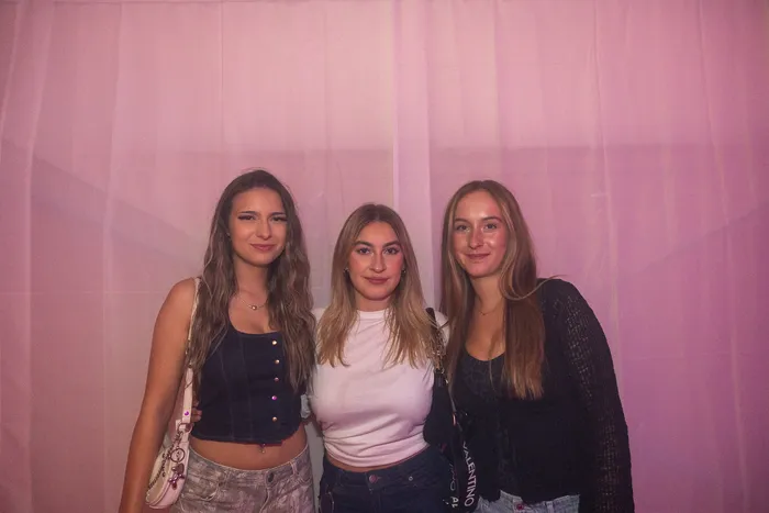 Drei junge Frauen posieren lächelnd vor einem rosa Vorhang bei der Q-Stall-Party.