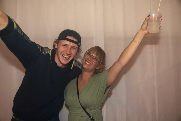 Ein lachender Mann mit schwarzer Kappe und eine Frau mit Brille und grünem Kleid posieren mit erhobenen Armen vor einem hellen Vorhang.