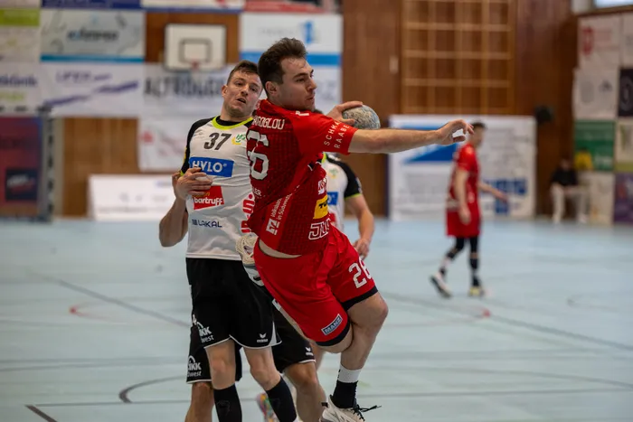 Handballspieler in roten Trikots springen während eines Spiels in einer Sporthalle. Ein Spieler wirft den Ball, während ein Gegenspieler ihn verteidigt.