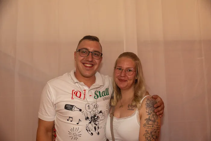Ein lächelndes Paar posiert vor einem hellen Vorhang; der Mann trägt ein weißes Poloshirt und die Frau ein weißes Top mit Tätowierungen am Oberarm.