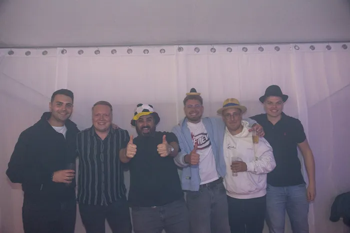 Sechs Männer posieren lächelnd vor einem weißen Vorhang und halten teilweise Getränke in den Händen.