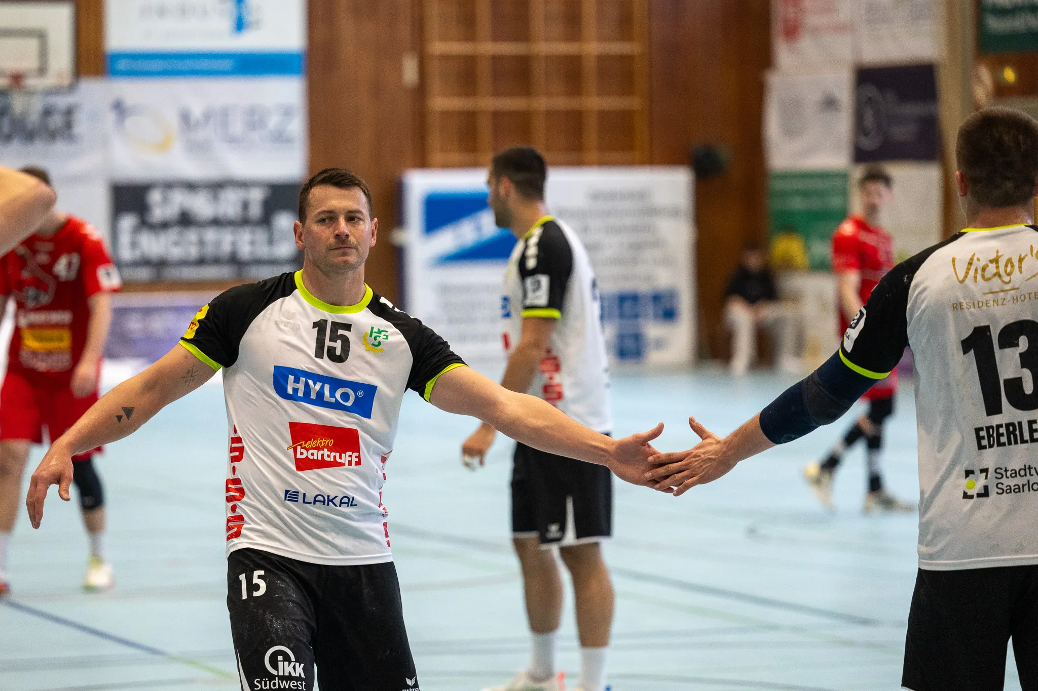 Handballspieler mit Trikotnummer 15 gestikuliert während des Spiels, im Hintergrund weitere Spieler und Zuschauerränge.