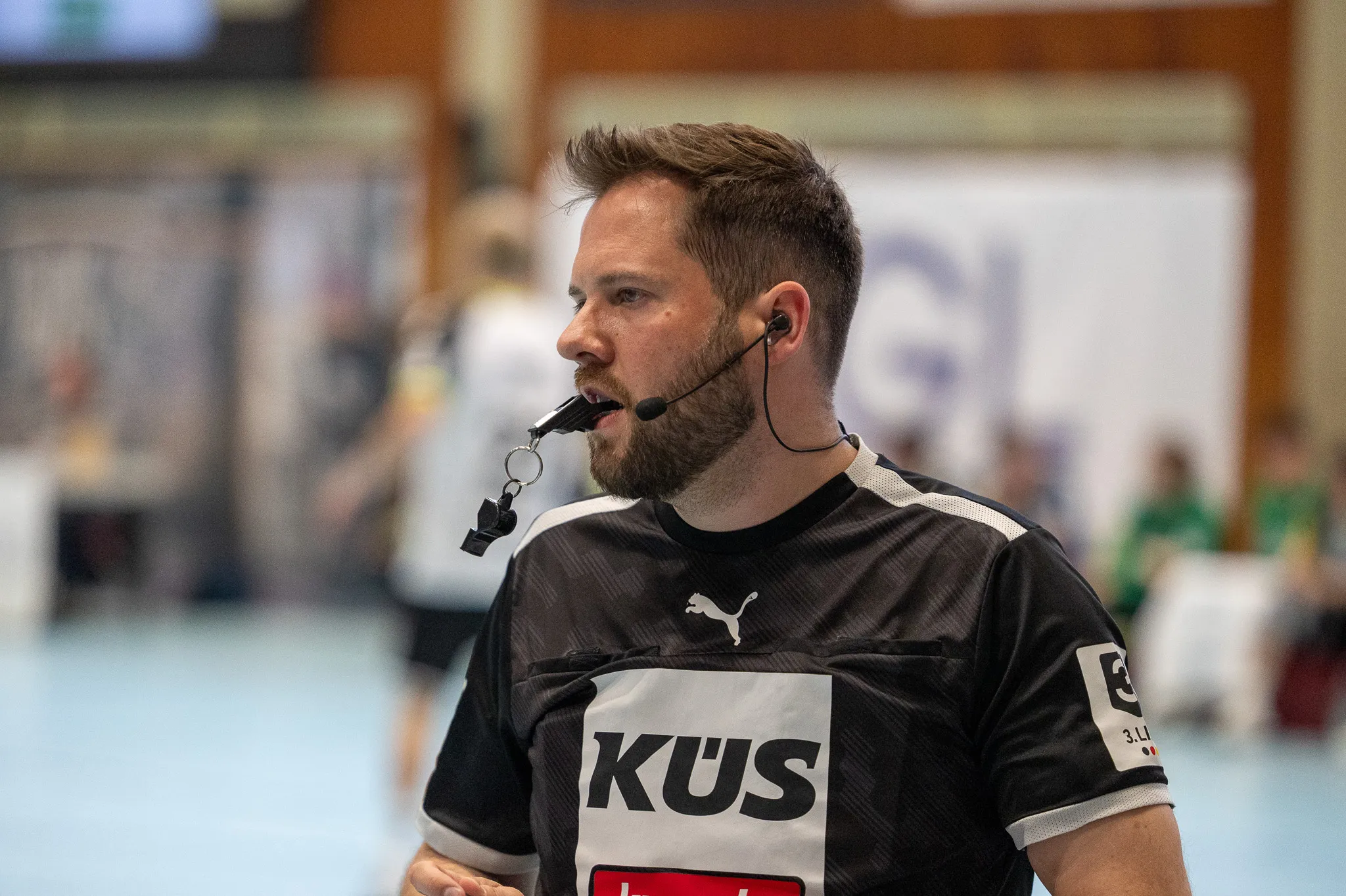 Handballschiedsrichter mit Bart und Headset pfeift während eines Spiels, trägt ein Trikot mit dem Sponsor-Logo KUS.