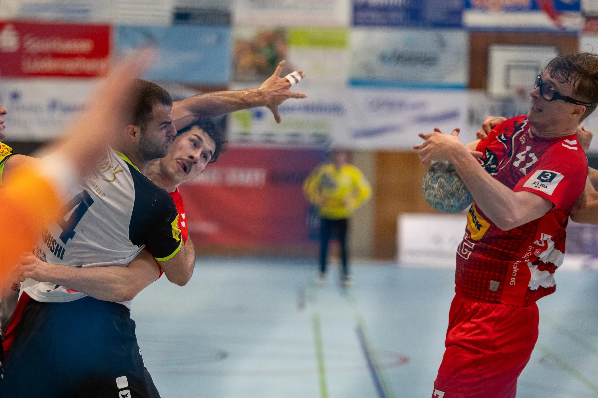 Zwei Handballspieler kämpfen um den Ball: Ein Spieler in Rot und Schwarz versucht, den Ball zu erobern, während ein Spieler in Rot und Weiß ihn verteidigt.