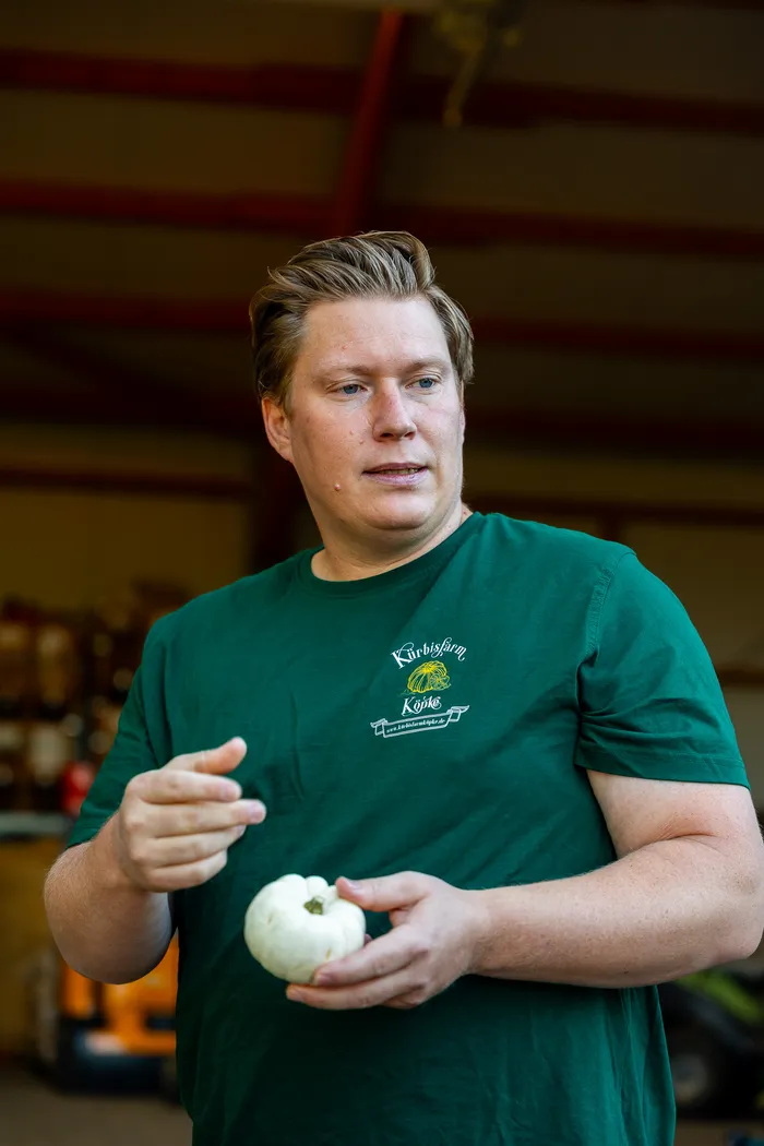 Ein Mann in einem grünen T-Shirt mit der Aufschrift „Kürbisfarm Köpke“ hält einen kleinen weißen Kürbis in den Händen.