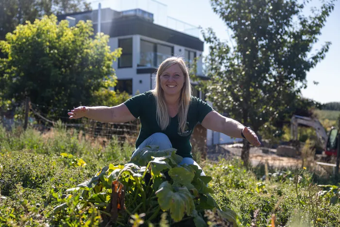 Eine lächelnde Frau kniet mit ausgebreiteten Armen vor einer großen grünen Pflanze auf einem Feld vor einem modernen weißen Haus.