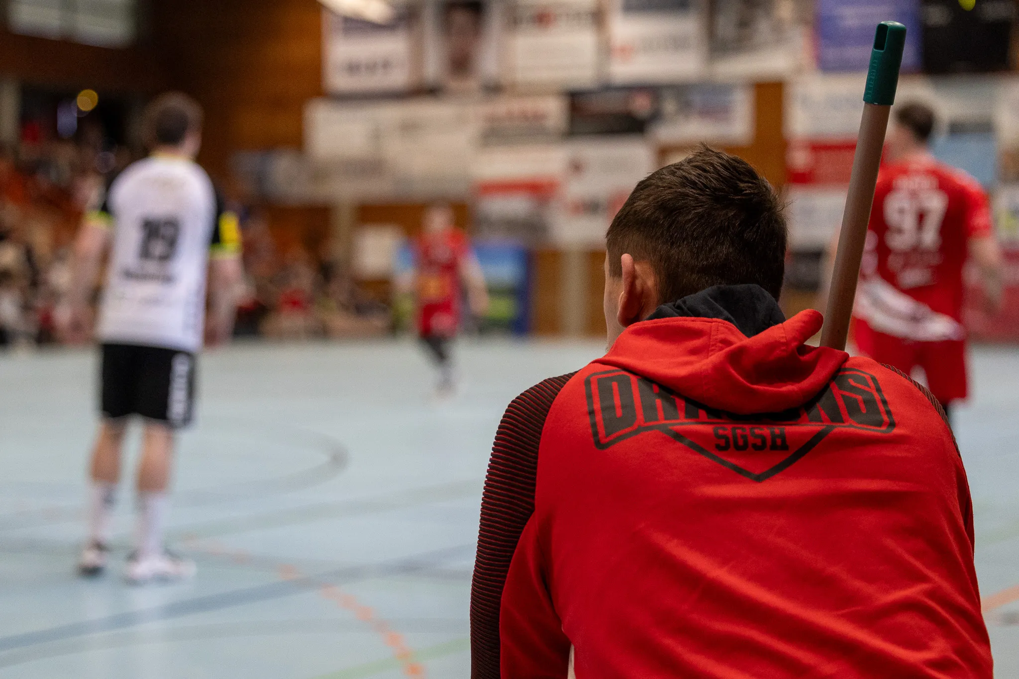 Rückansicht einer Person mit rotem Hoodie und Floorballschläger während eines Spiels. Im Hintergrund sind weitere Spieler und Zuschauer in einer Sporthalle zu sehen.