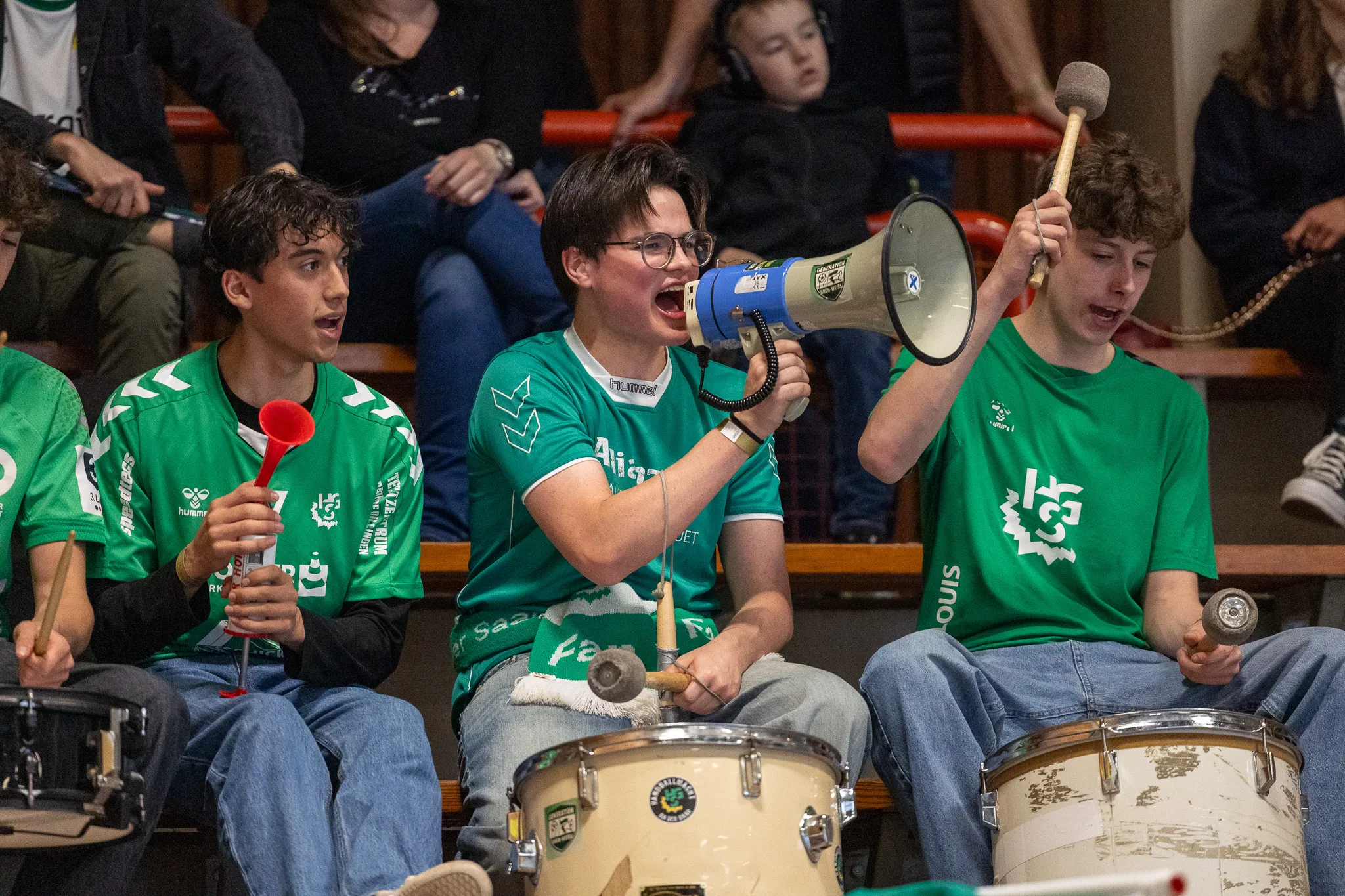 Mehrere junge Fans in grünen Trikots feuern bei einem Handballspiel an. Ein Fan hält einen Megafon in die Höhe, während andere mit Trommeln und Rasseln Lärm machen.