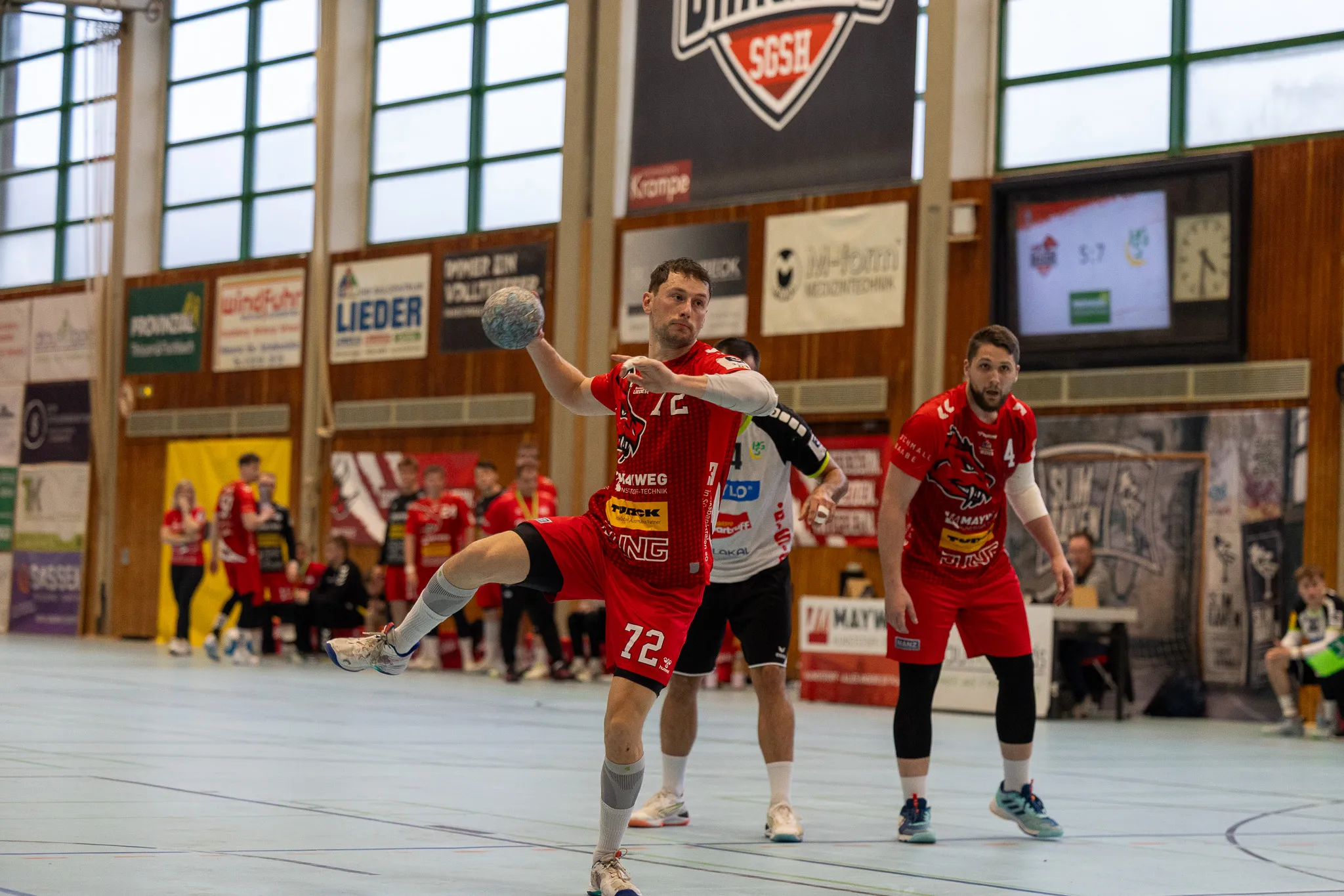 Handballspieler in roten Trikots mit der Nummer 72 springt zum Wurf, während ein Gegenspieler ihn verteidigt. Im Hintergrund die Halle und weitere Spieler.